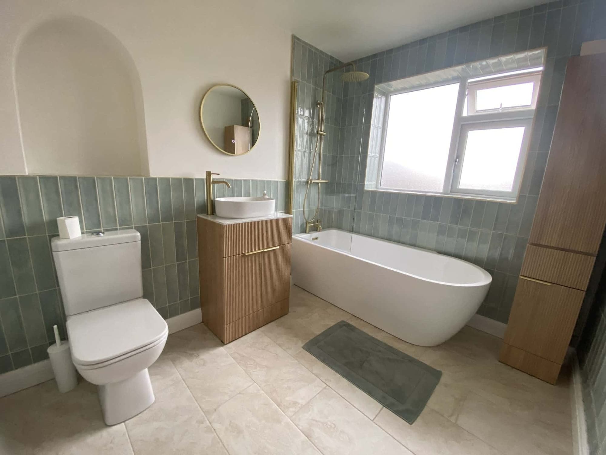 The same bathroom after renovation, with sage green vertical tiles, brushed brass fittings, an oak-effect vanity, a round mirror in an arched alcove and a freestanding bath