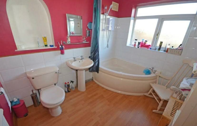 Dated bathroom before renovation, with bright pink walls, a corner bath, pedestal basin and wood-effect flooring