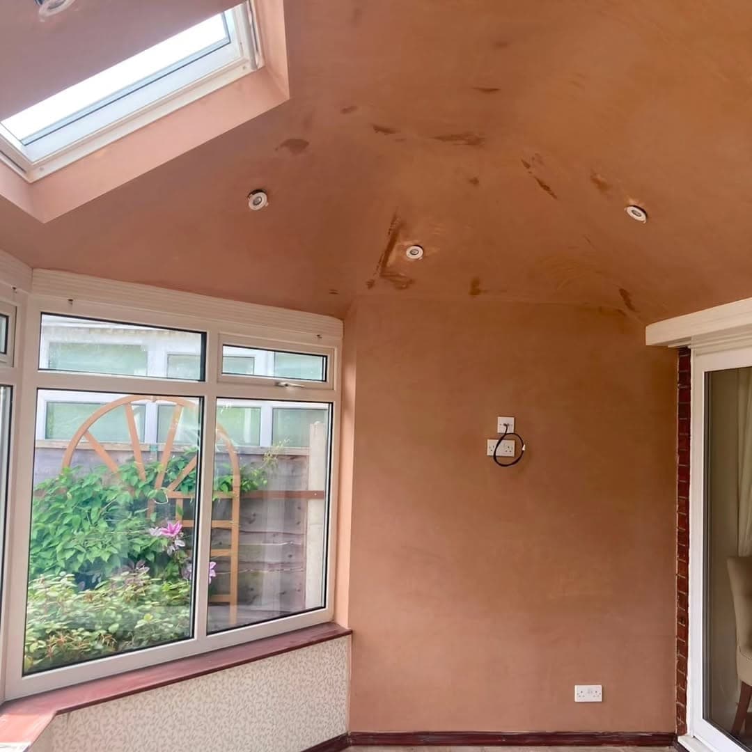 Newly plastered conservatory ceiling with recessed downlights fitted, above a window looking onto a garden