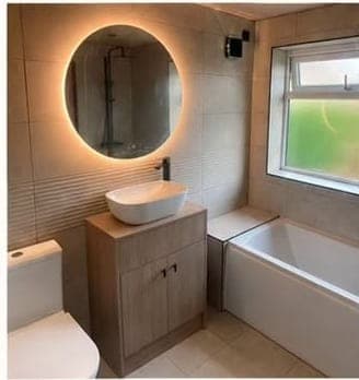 Warm neutral bathroom with a backlit circular mirror above an oak-effect vanity, beside a panelled bath