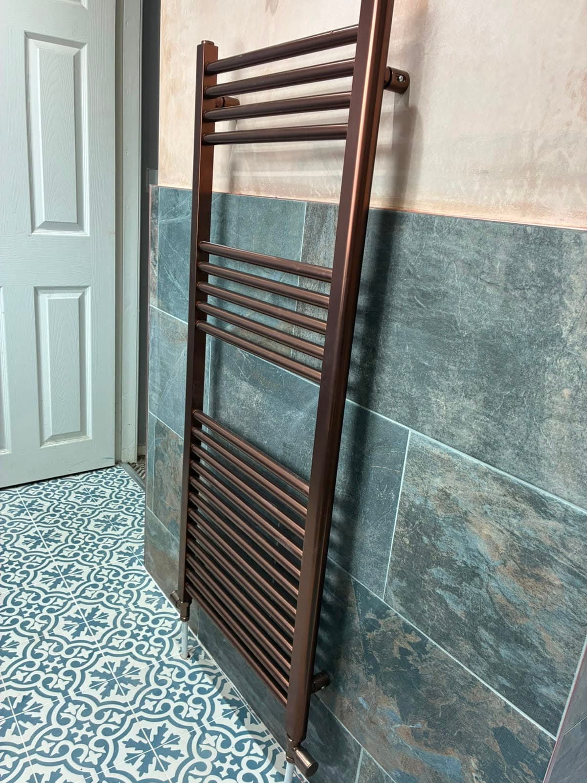 Copper-finish ladder radiator fitted on a slate-effect tiled wall above a blue patterned tiled floor