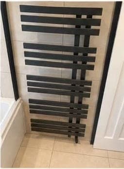 Matt black flat-bar ladder radiator fitted on a beige-tiled bathroom wall above the end of a bath