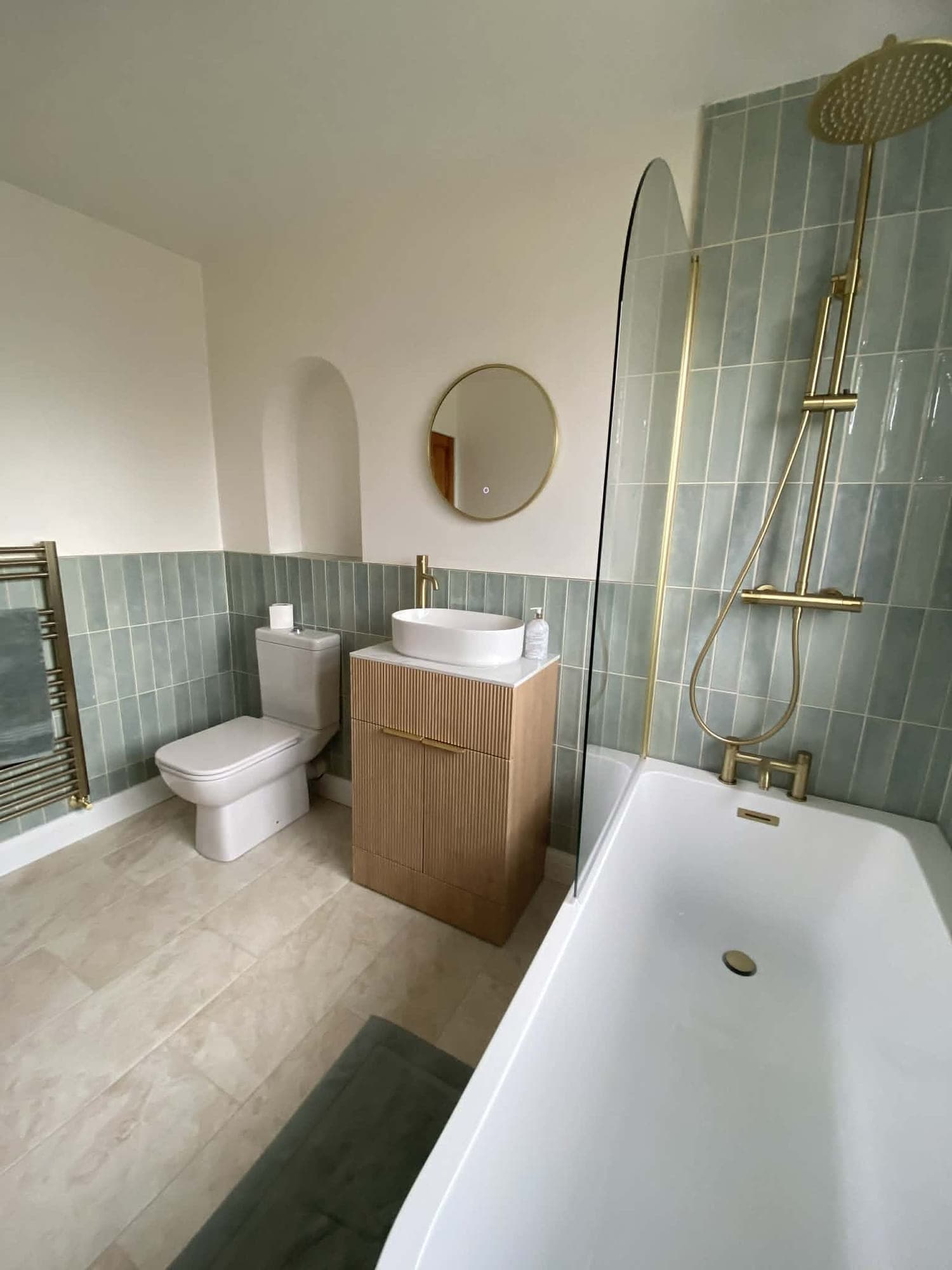 Sage green tiled bathroom with a brushed brass rainfall shower over the bath, a round mirror and a heated towel rail