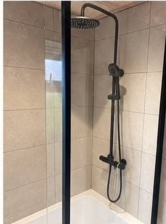 Matt black rainfall shower and handset over a white panelled bath, against large-format beige tiles behind a black-framed screen