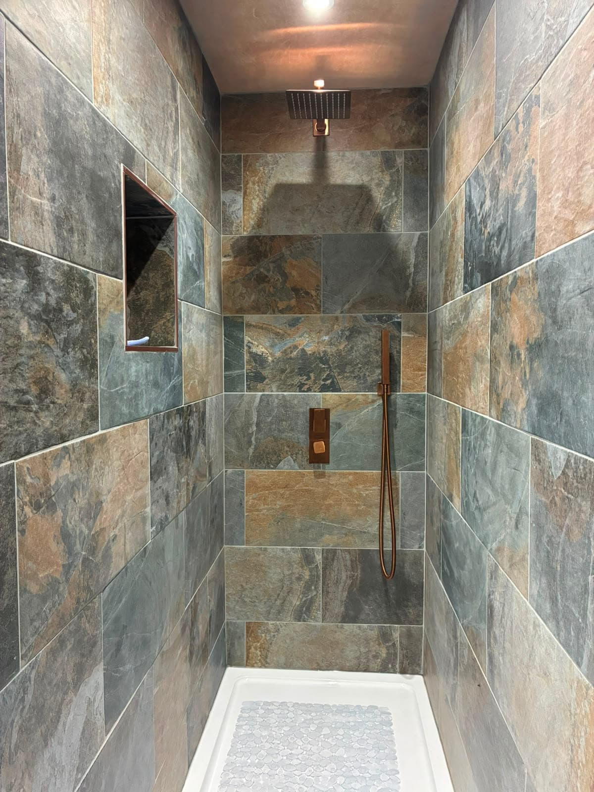 Walk-in shower tiled in slate-effect stone with a recessed niche and a copper rainfall shower head