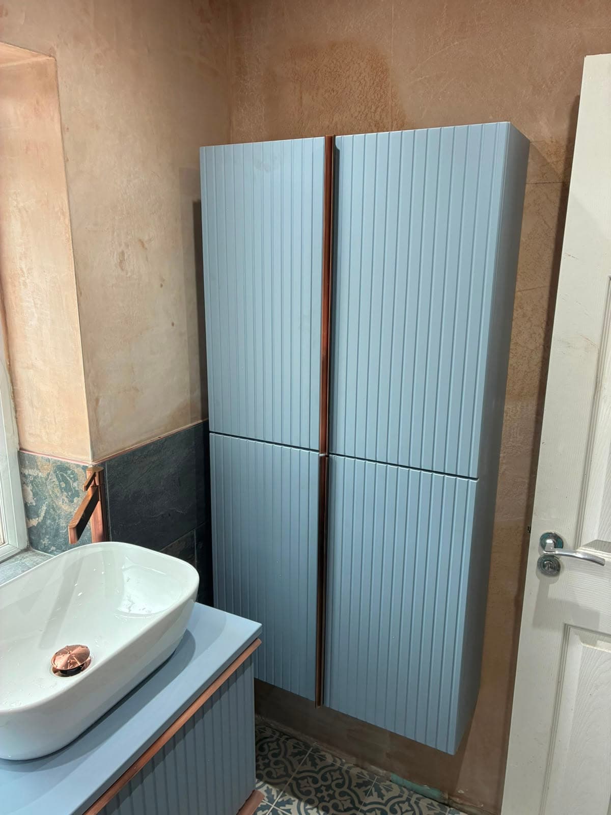 Blue-grey fluted tall storage cabinet beside a countertop basin in a slate-tiled bathroom