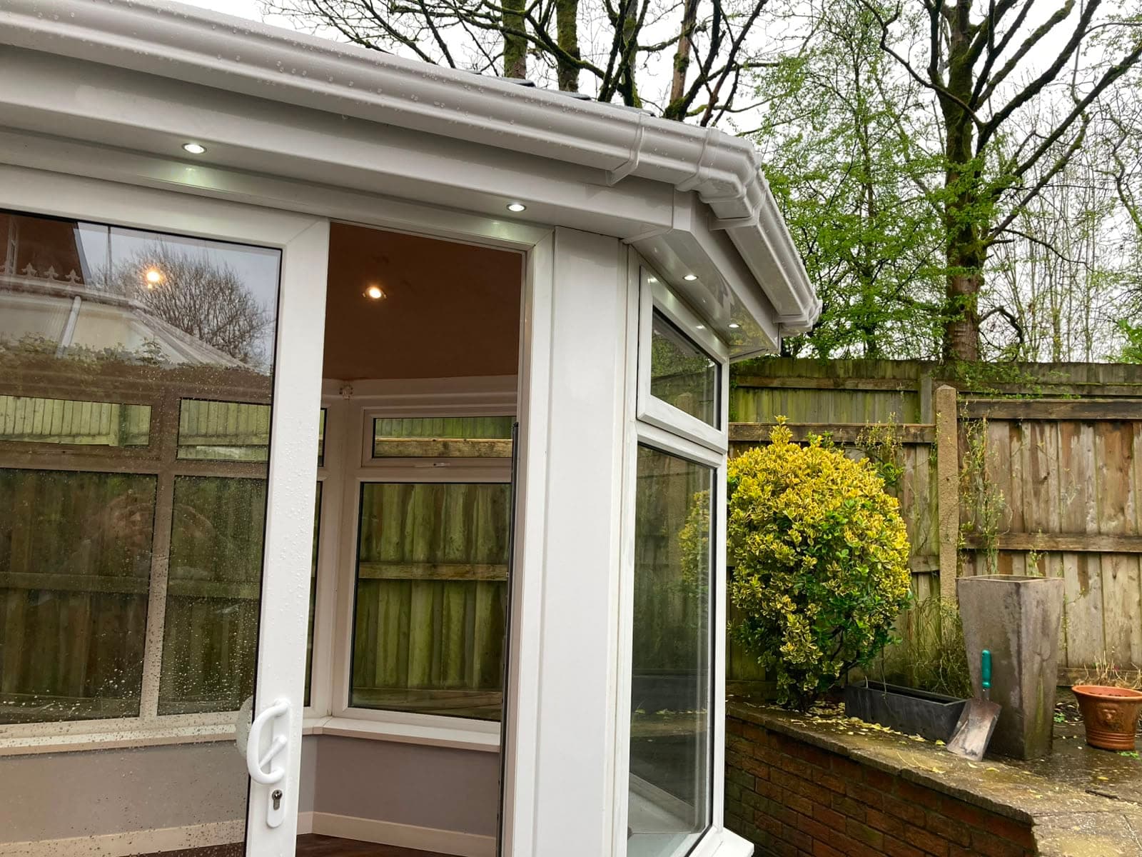 Interior of a converted conservatory with white frames, a plastered ceiling and downlights, looking onto the garden